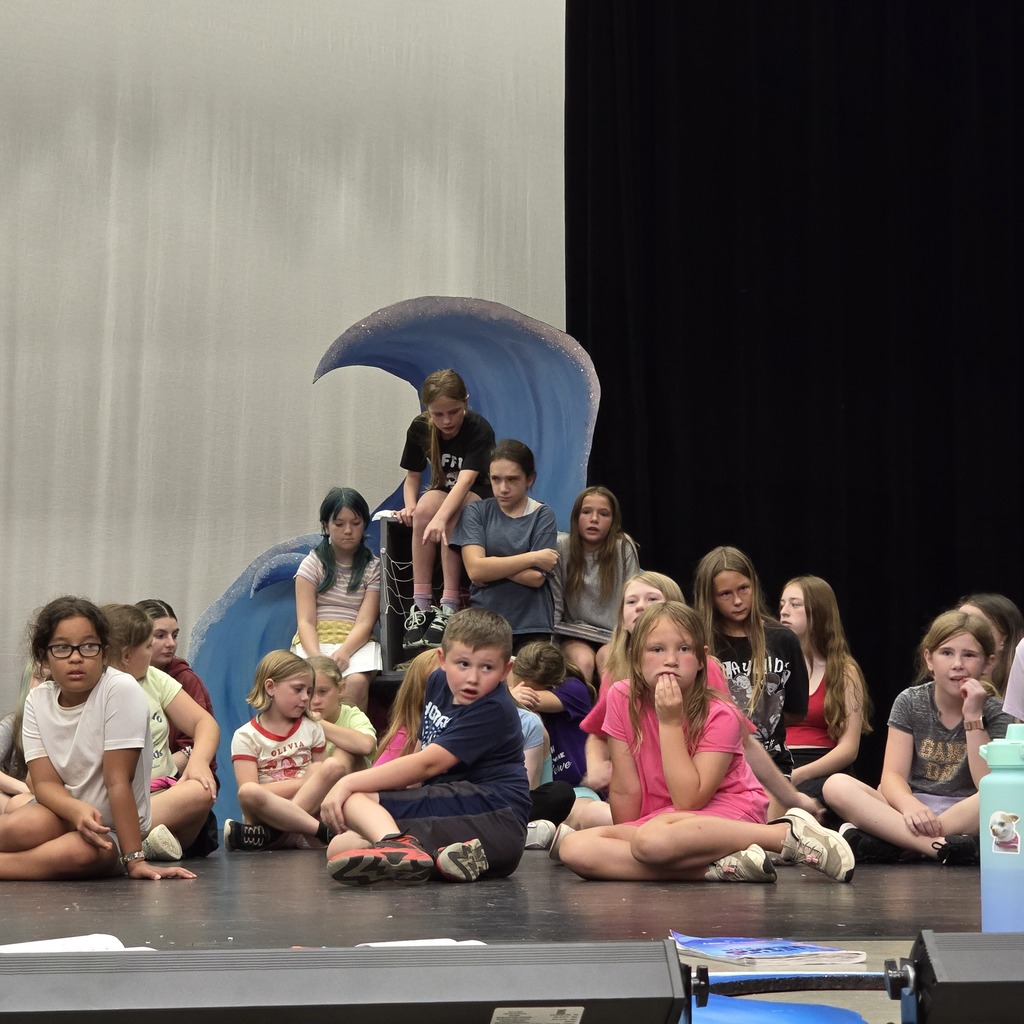 Students sit on stage with a blue wave behind them.