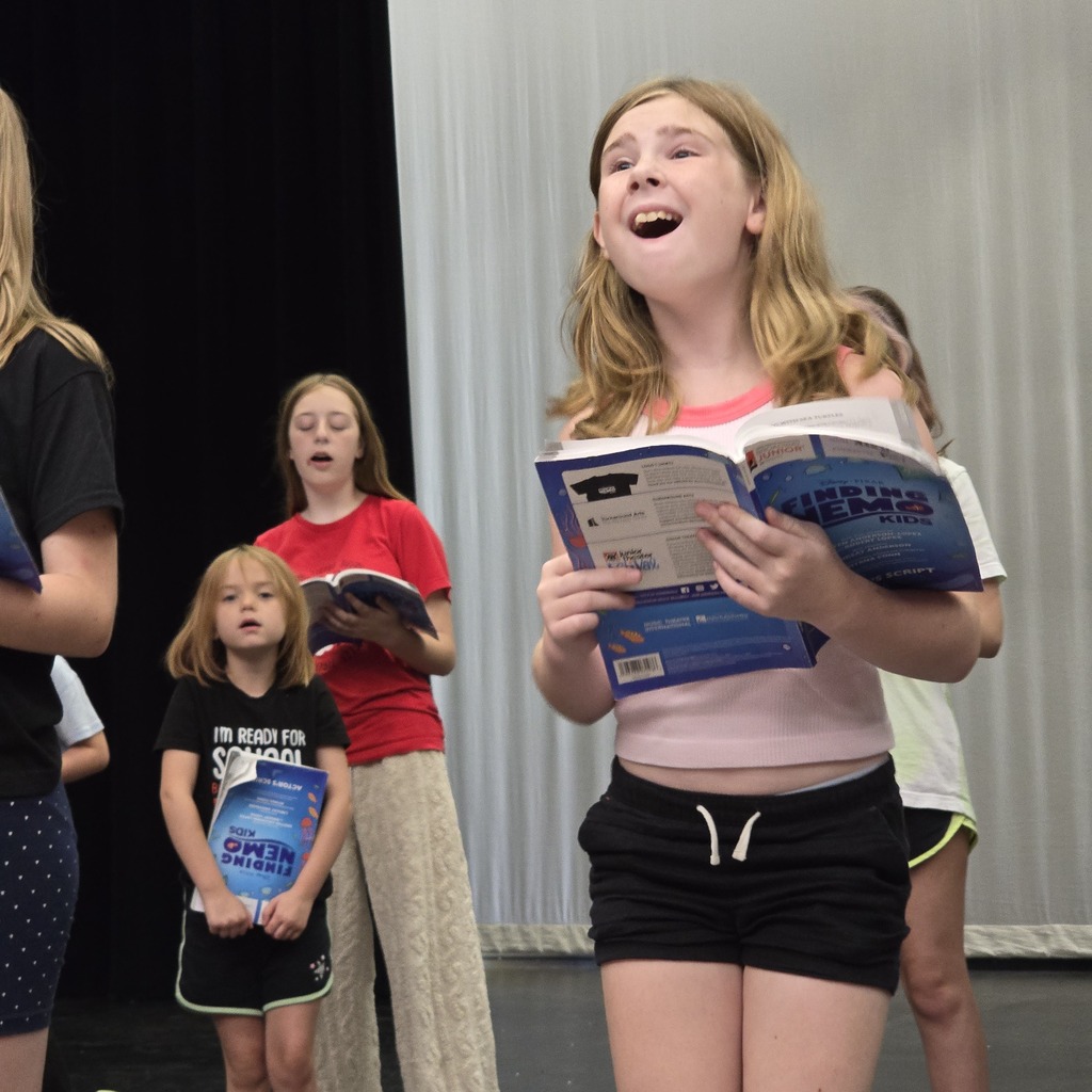 A group of children singing, holding books, with white fabric curtains in the background.