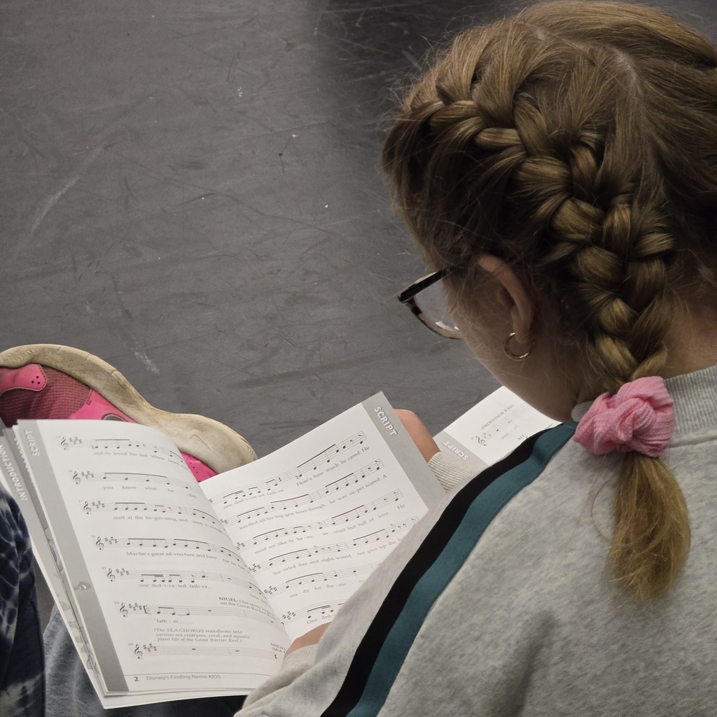 A person reads a music book on a stage floor, wearing glasses and a braid with a pink flower.