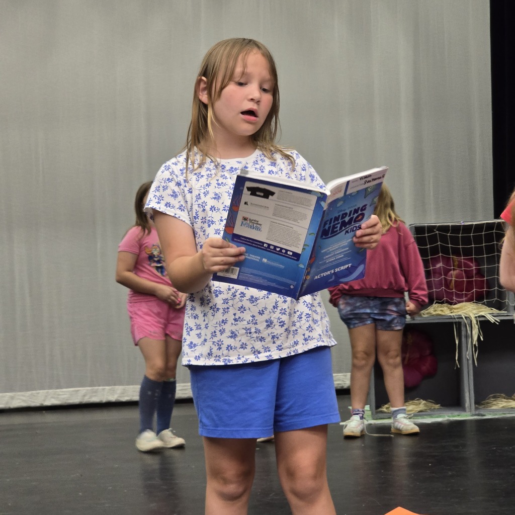 A girl reads a book on stage with two other girls behind her, all wearing casual clothes.