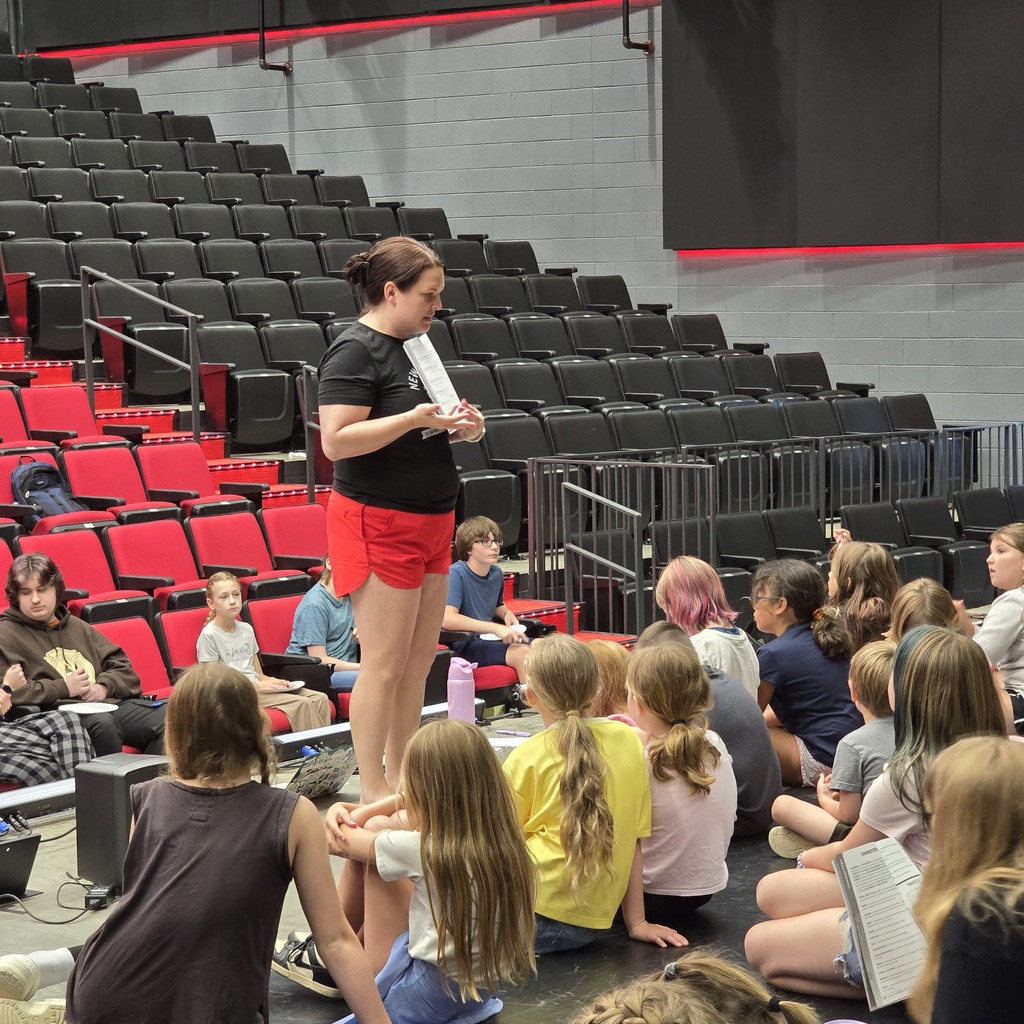 A woman stands holding a paper in an auditorium; children sit in chairs, facing her, and appear to be listening.