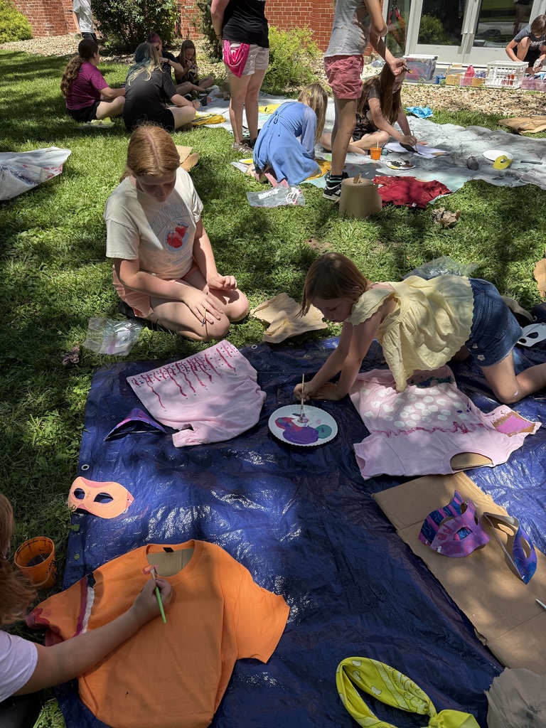 Kids sit on a grassy field, engaging in a painting activity on a blue tarp. Various art supplies are scattered around them.