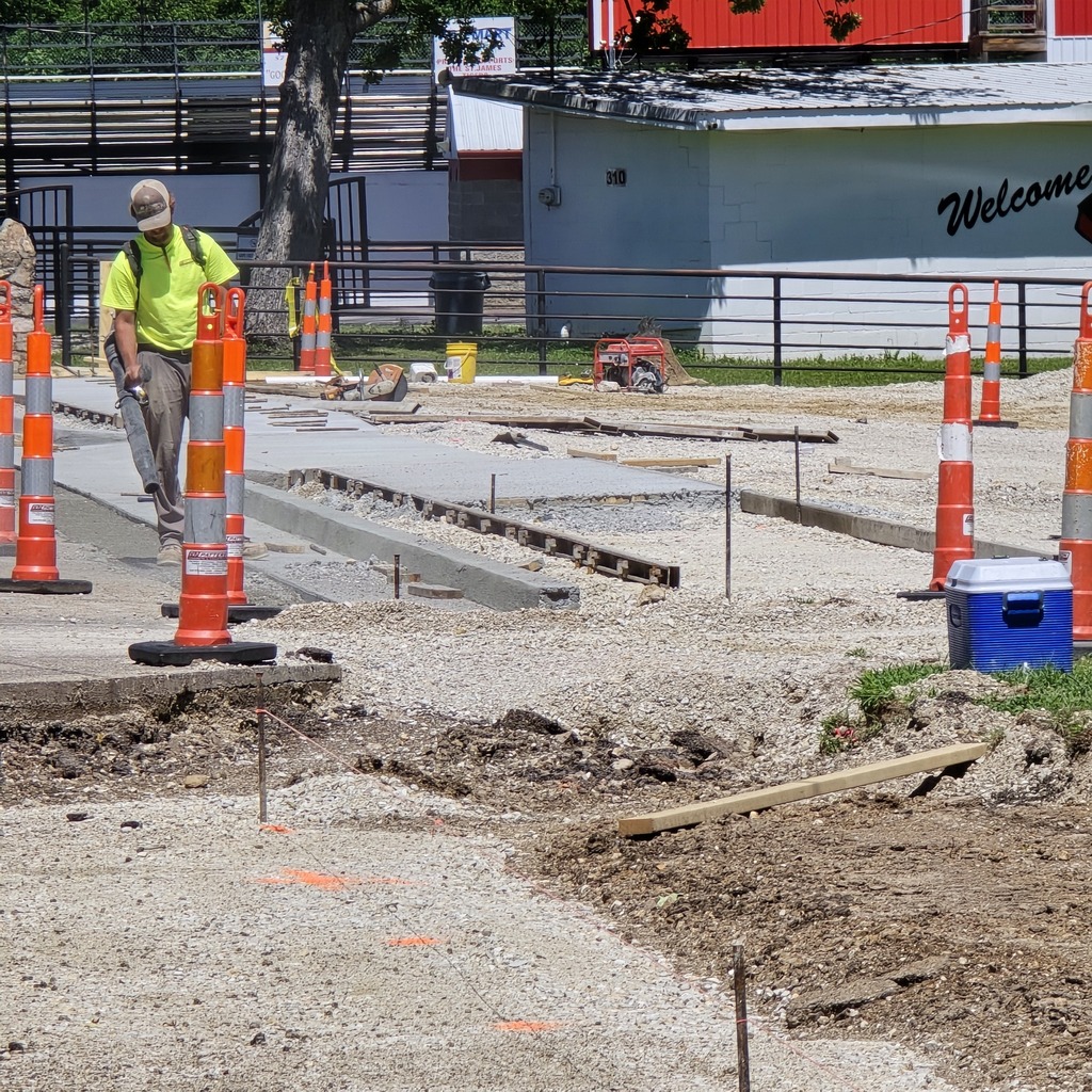 A man in a yellow shirt and hat walks past a construction site with orange and white traffic cones.