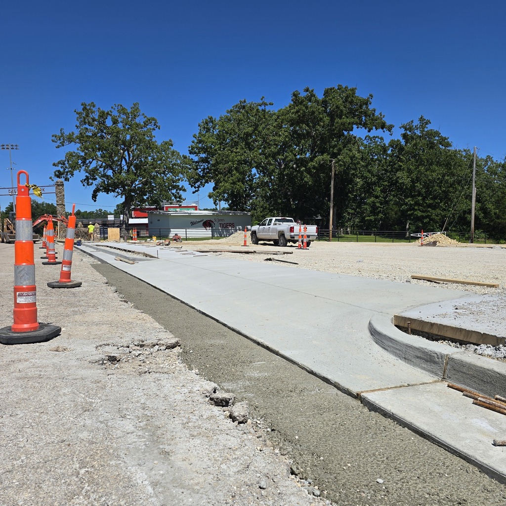 Newly poured concrete pathway under construction. Trees and greenery visible in the background.