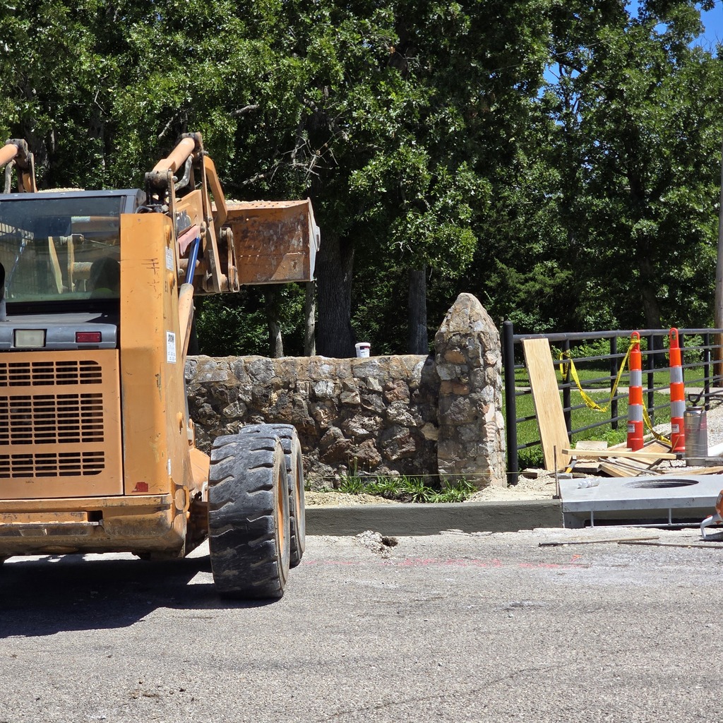 A large orange construction vehicle with a raised bucket on a paved area. Behind it, a stone wall, trees, and construction barriers.