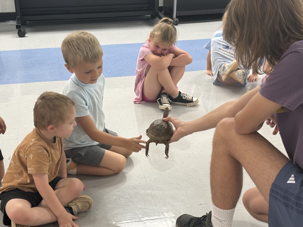 A man holds a turtle while young children sit around him, watching. They are in a classroom.