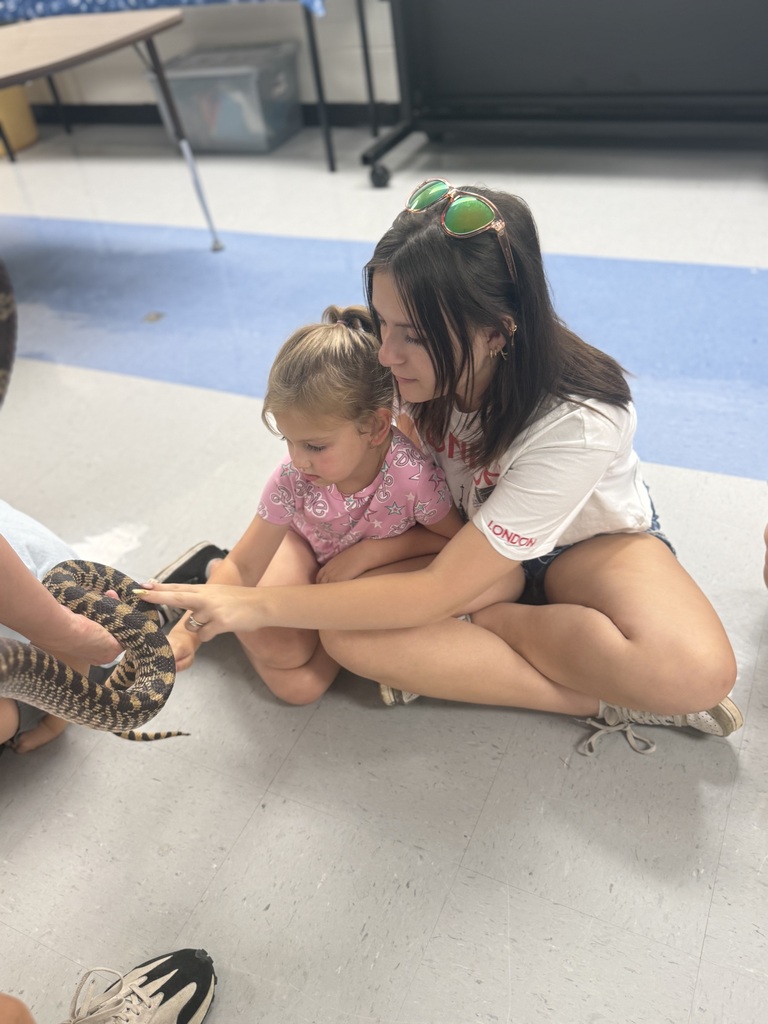 A woman holds a snake with a child in a classroom setting; both are sitting on the floor.