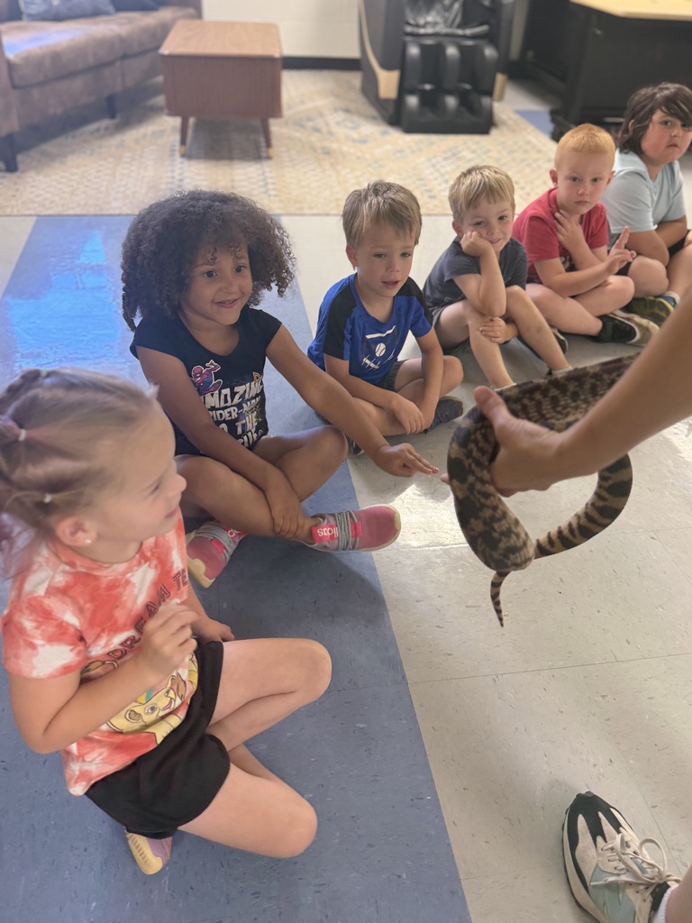 Five children sit on the floor, watching someone hold a snake. They wear casual clothing and sit on a blue mat.