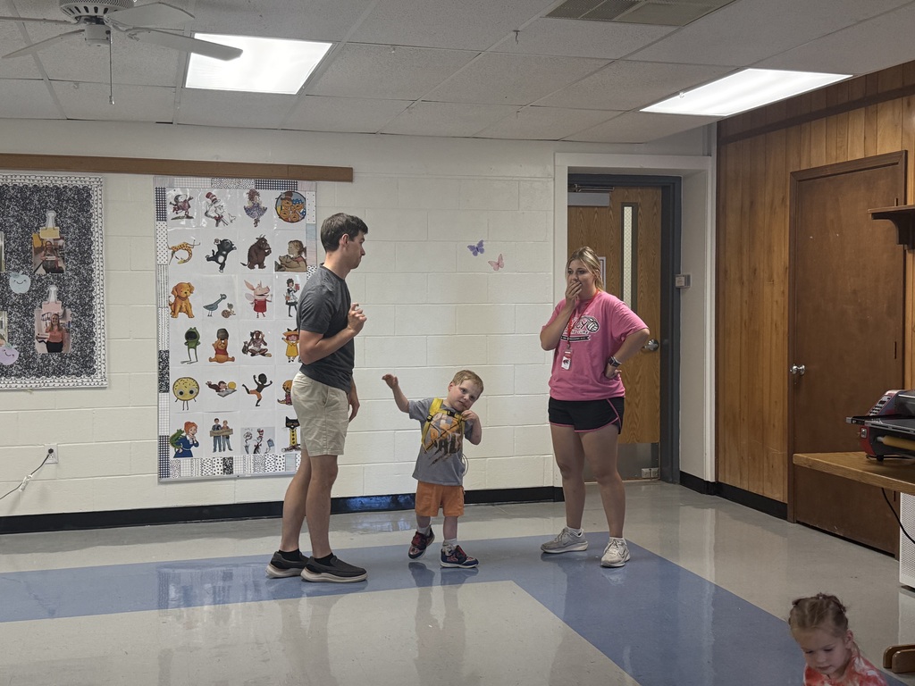 Three people in a room, one with a child. The child holds the adult's hand. A wall has various pictures.