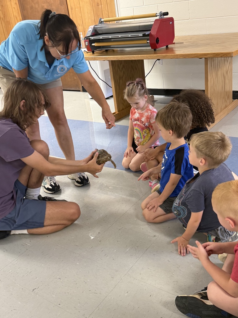 Several children sit in a circle as a woman holds a small reptile. Behind them is a wooden table.
