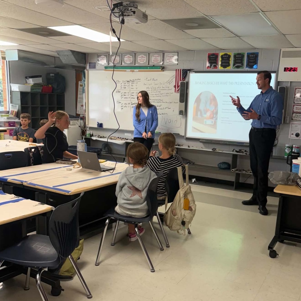 A classroom with students seated, a teacher presenting on a projector screen, a whiteboard, and a projector on the ceiling.