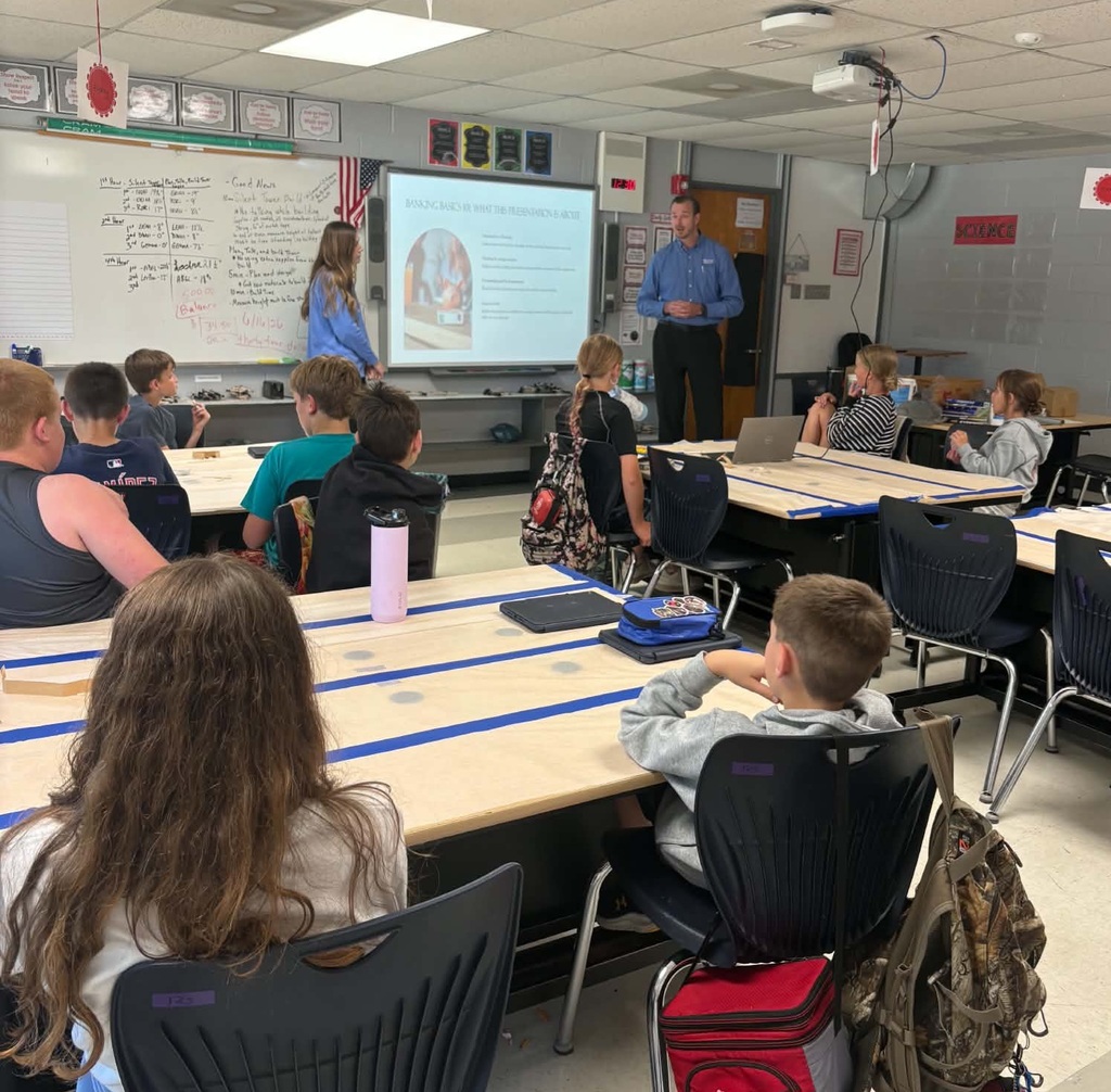 A classroom with a teacher standing in front of a whiteboard. Students sit at desks with backpacks.