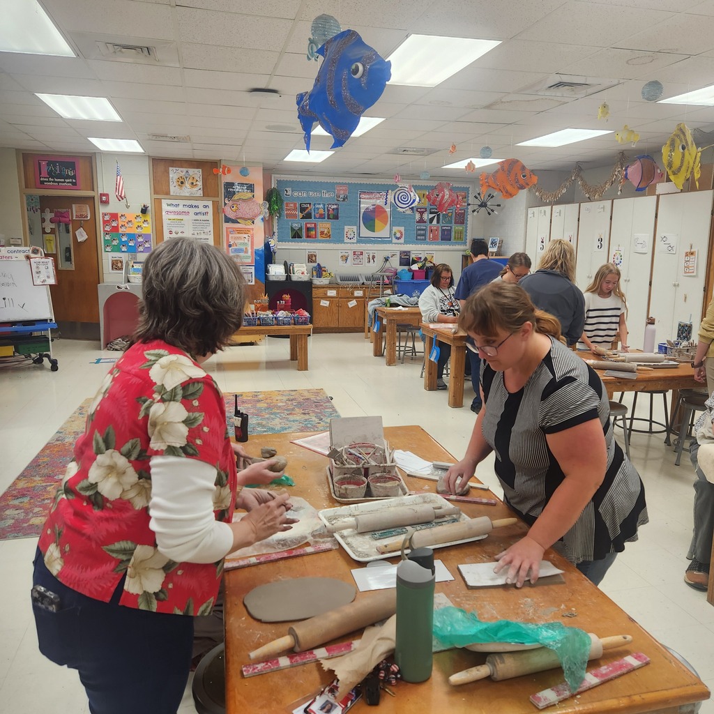 Two women stand at a table with rolled dough and other items. They are in a room with other people, a rug, and cabinets.