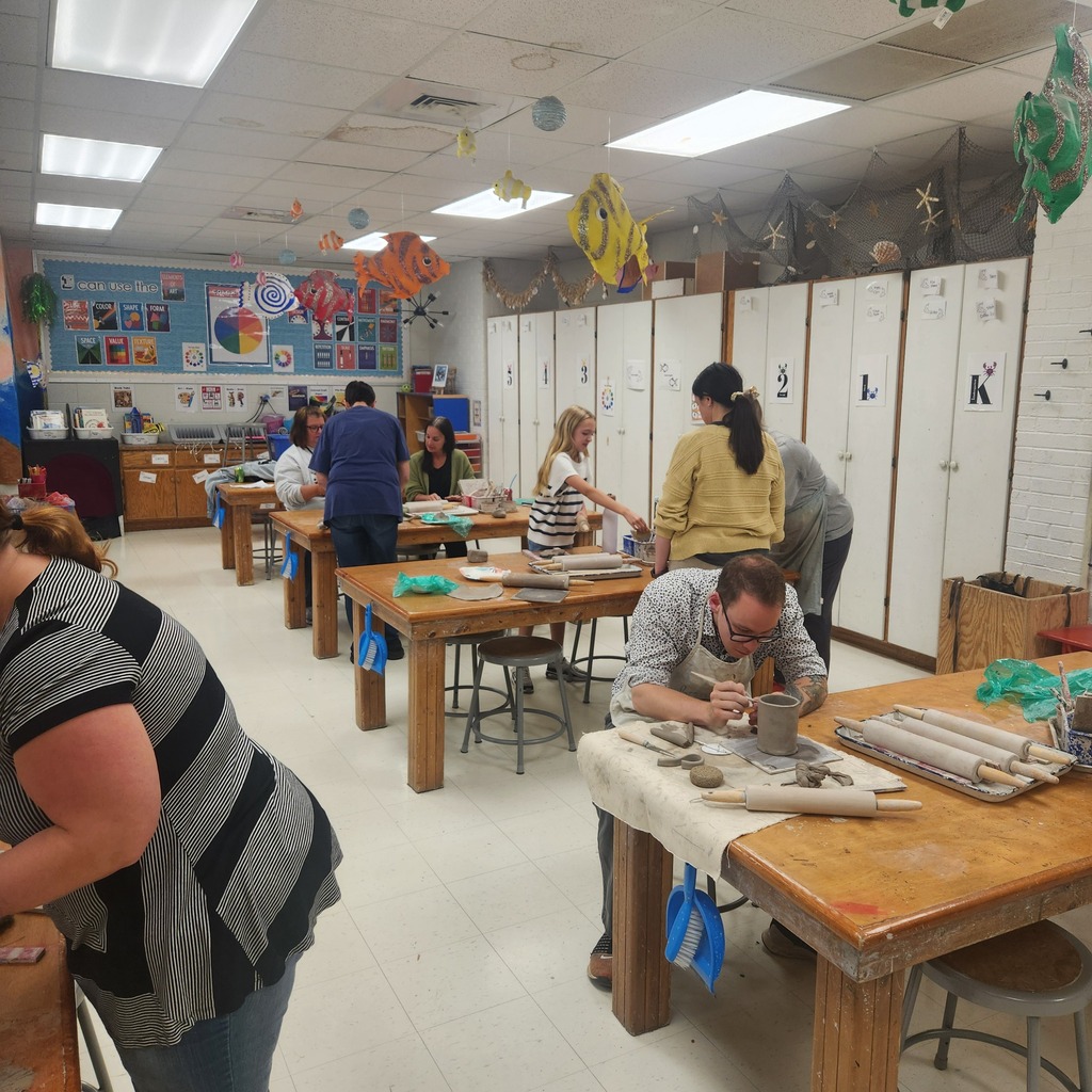 A group of people working on pottery in a room with long wooden tables and cabinets.