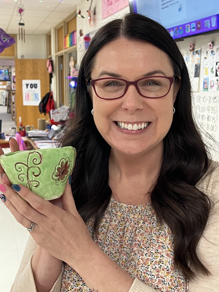 Woman with glasses holds green bowl with floral design, smiling in a classroom.