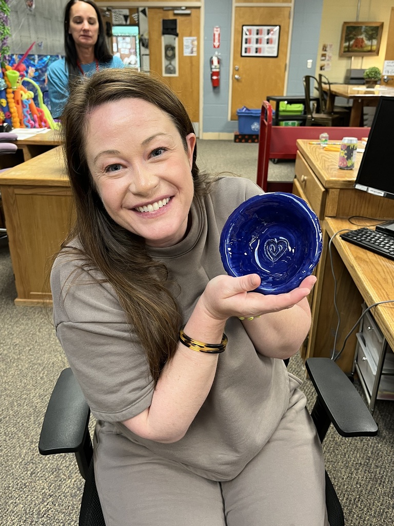 A woman in a gray shirt holds a blue ceramic bowl in an office with desks.