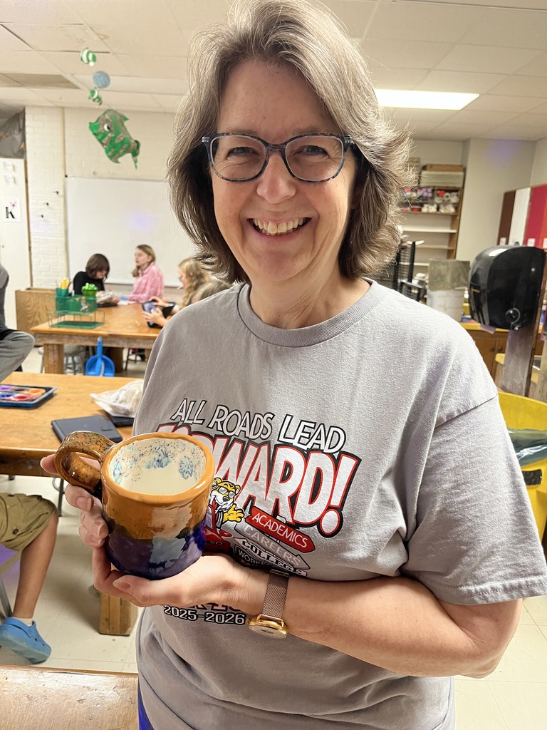 An adult woman wearing glasses and a gray t-shirt smiles while holding a ceramic mug. A classroom with tables and chairs is in the background.