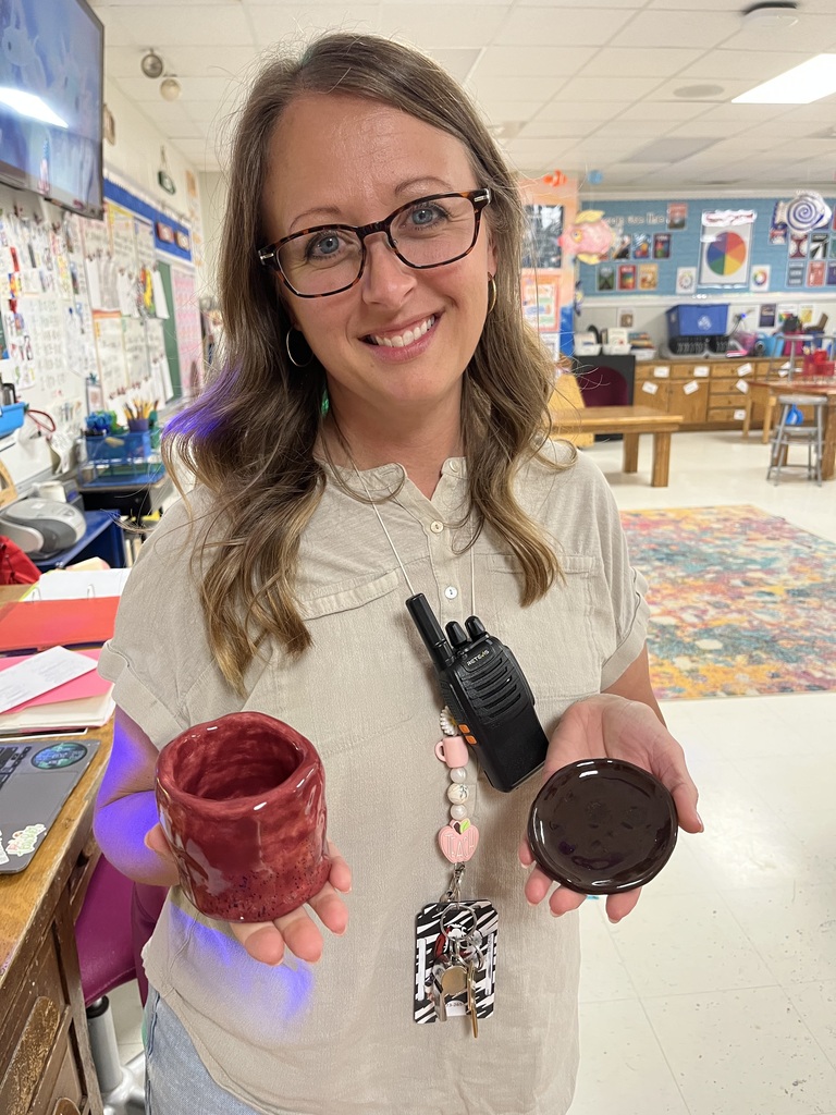 A woman with glasses and a necklace holds two red cups and a black walkie-talkie in a classroom.