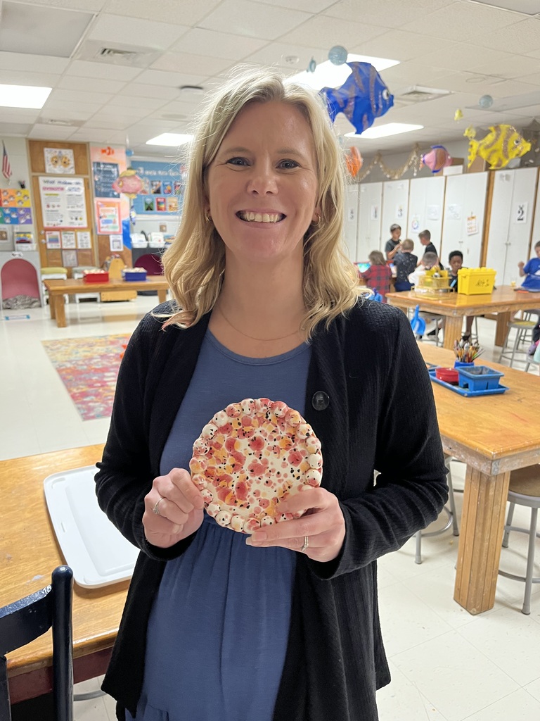 A woman holds a pizza in front of her stomach in a classroom. Desks and chairs are in the background.