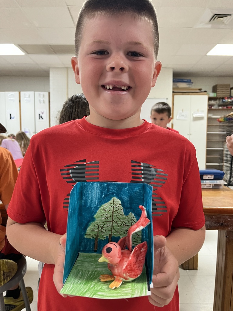 A boy shows a crafted paper scene of a red bird in a blue box.