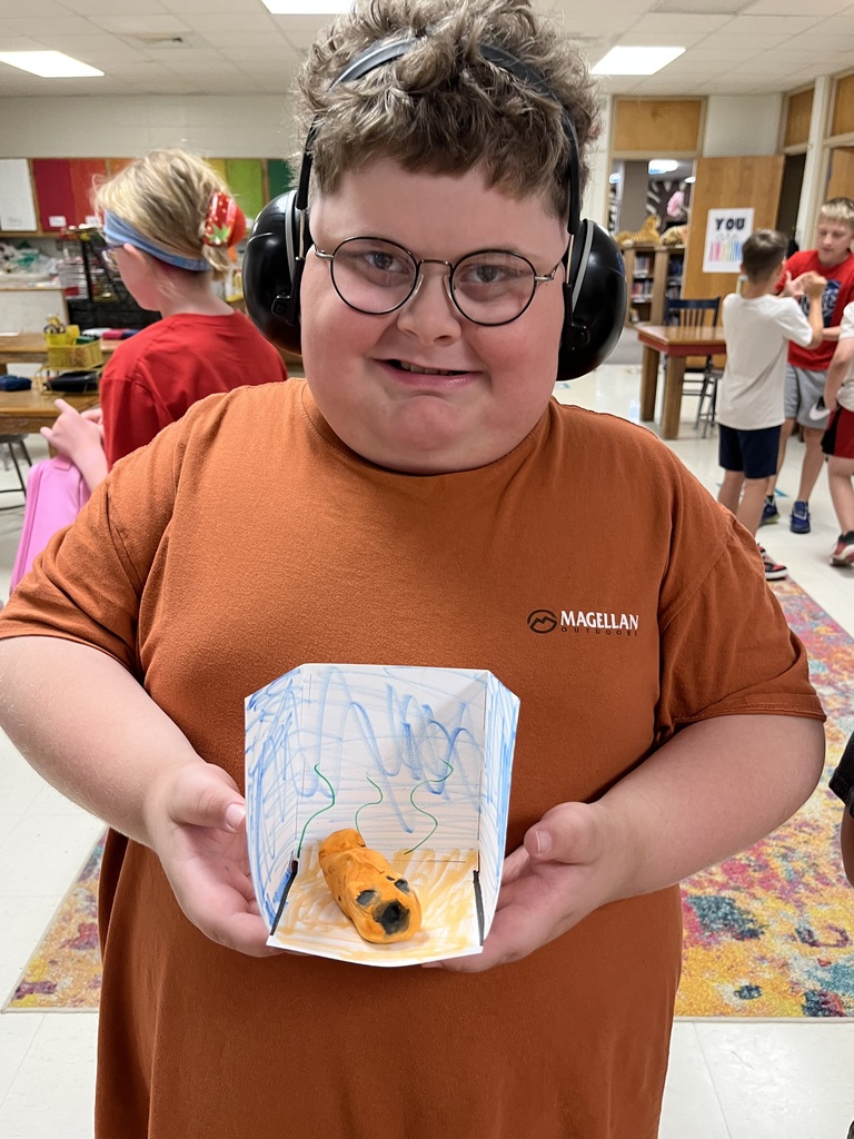 Person in brown shirt and headphones holds a paper model of a snake. Background shows a classroom with students.