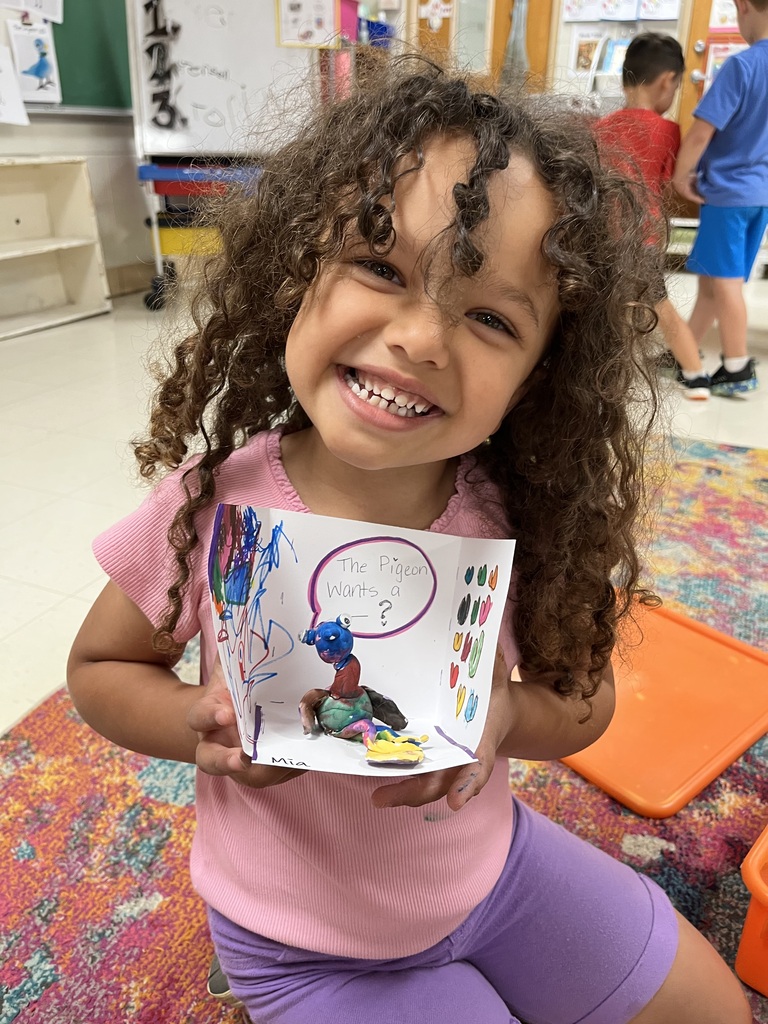 A young child with curly hair, wearing a pink shirt and purple shorts, holding a colorful card. Behind them is a classroom setting with a rug and other children.