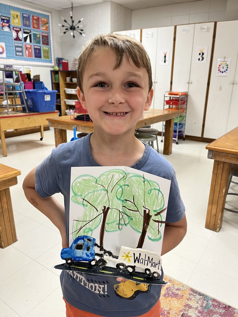 A child in a blue shirt and orange shorts holds up a drawing of a tree and toy cars.