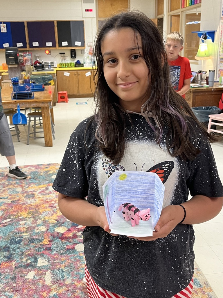 Girl in classroom holding a clear box with pink object inside, another student visible behind her.