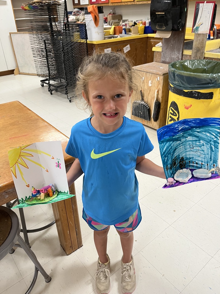 A child holds two colorful drawings, one with a sun and another with a landscape, in a room with a wooden table and a trash bin.