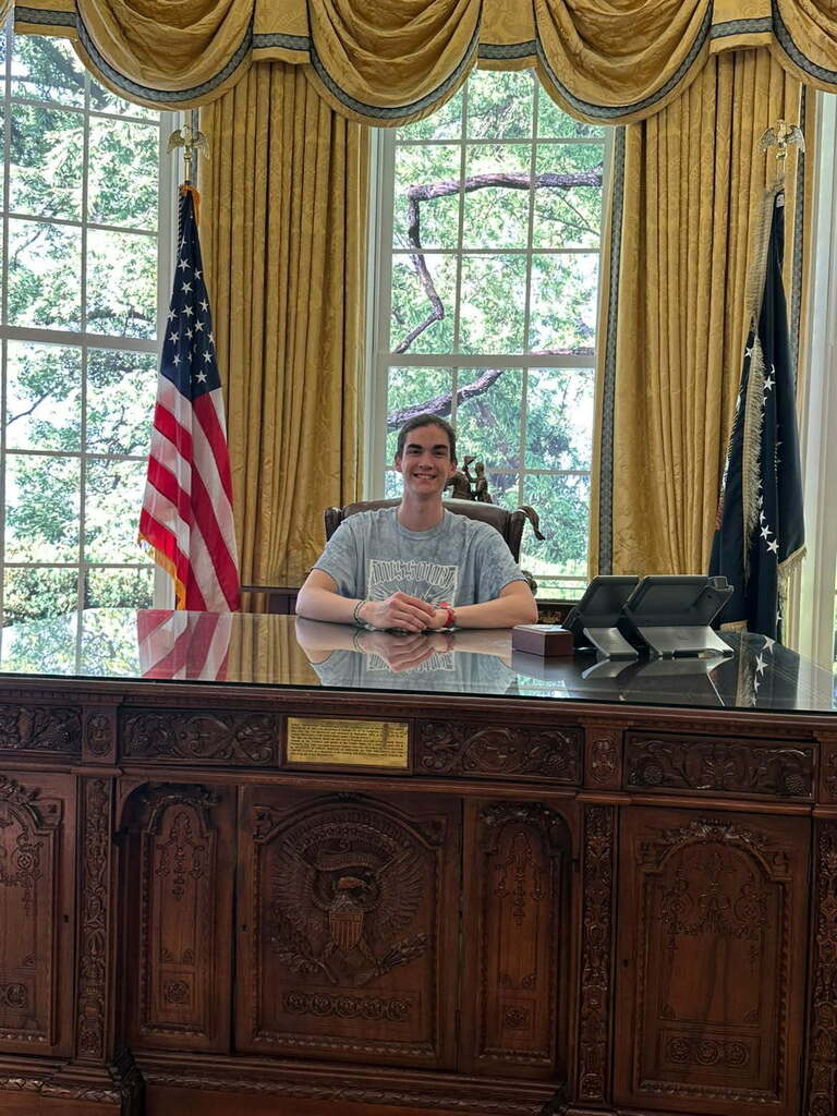 A person sits at a wooden desk with an oval plaque in a room with large windows.
