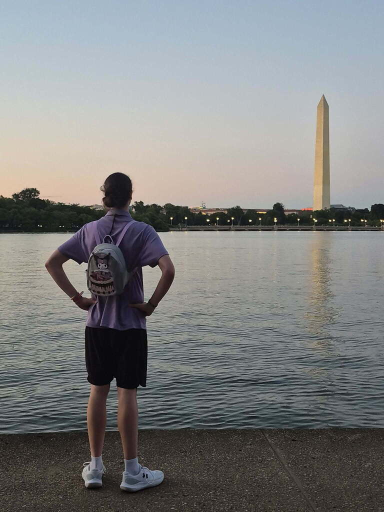 A man with a backpack stands by a lake, facing the Washington Monument. The sky is clear.