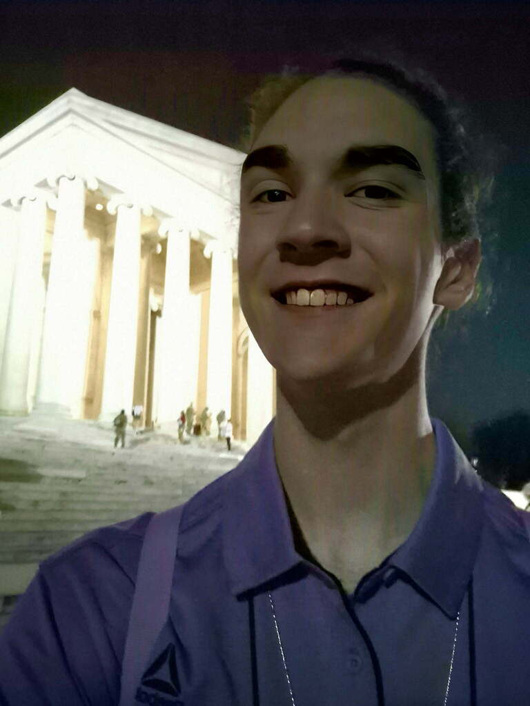 A smiling person stands before a historic building with multiple columns, under a night sky.
