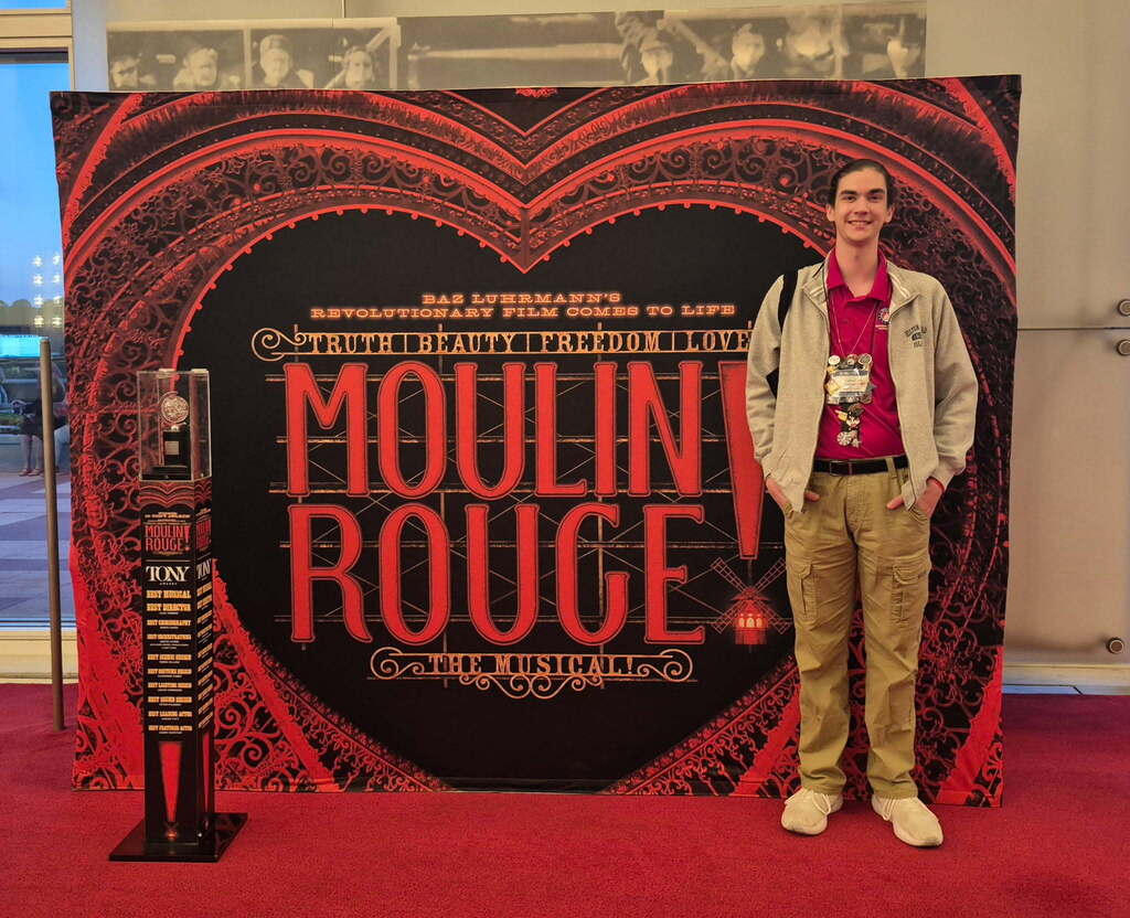 A person stands next to a Moulin Rouge display with a heart-shaped sign, wearing a red shirt and khaki pants.