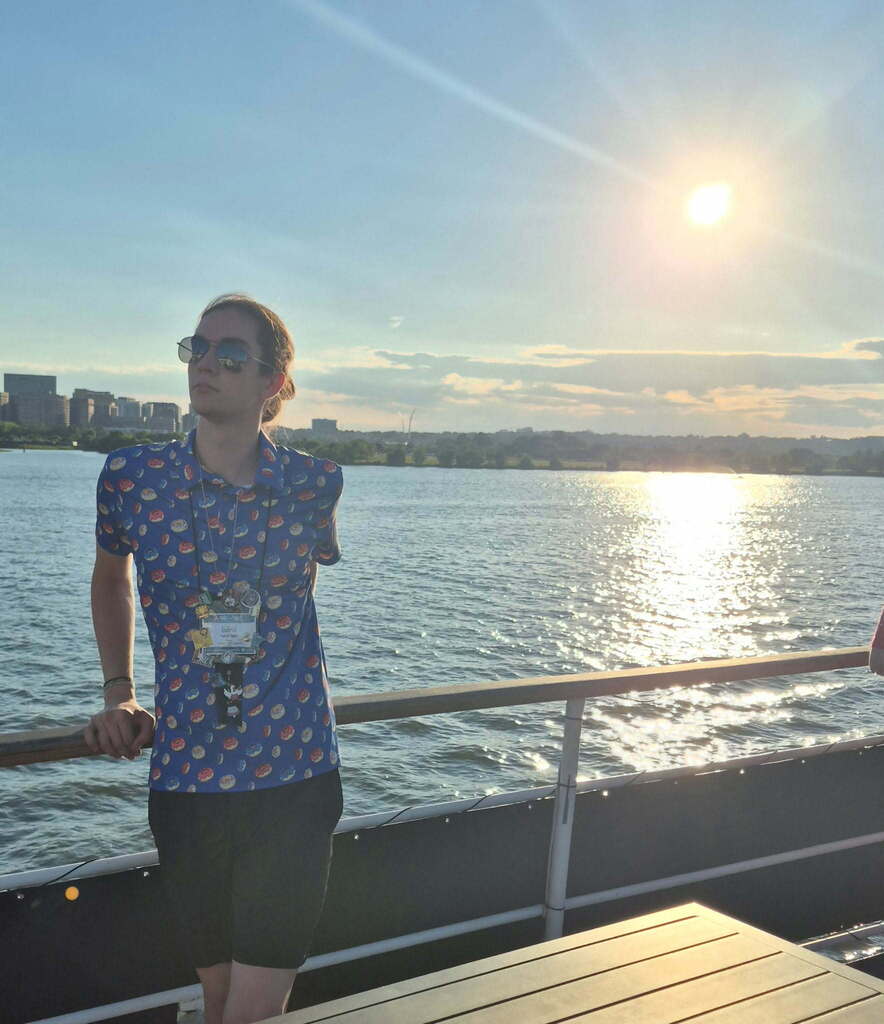 A person stands on a boat deck, looking at the sunset over water, wearing sunglasses and a patterned shirt.