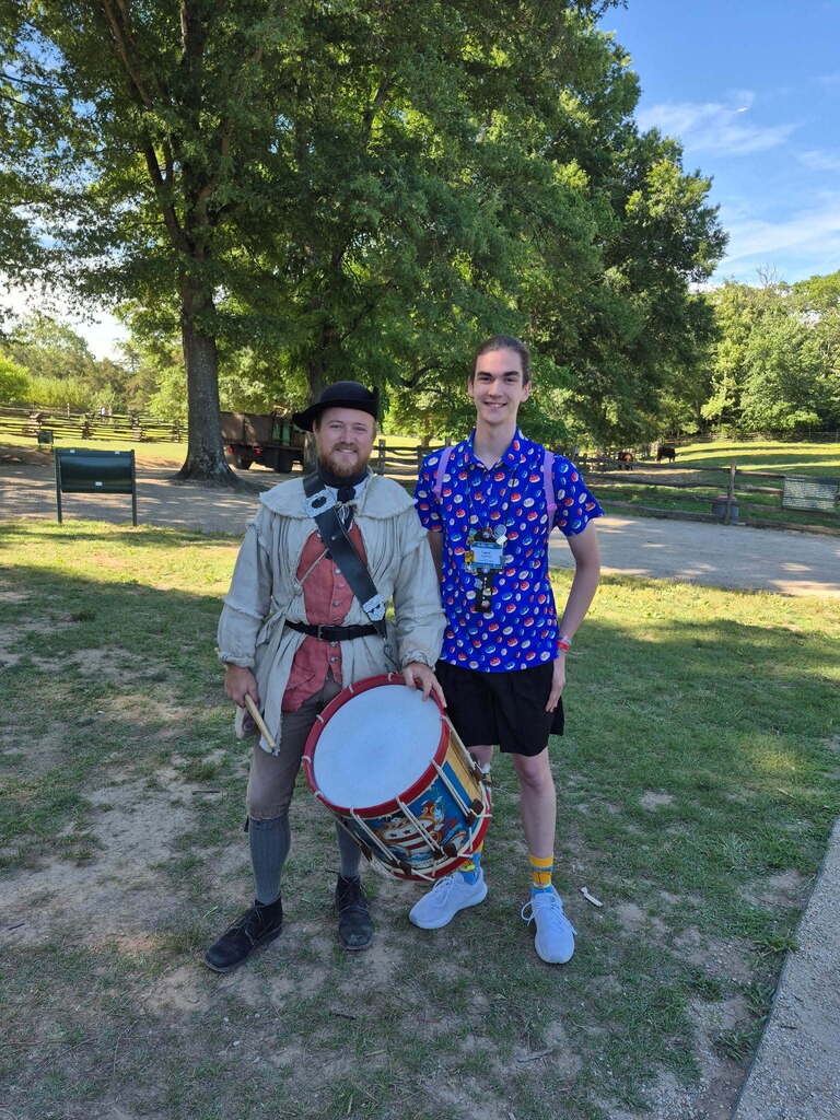 Two men stand outside on grass. One wears historical attire, holding a drum. The other wears casual clothes. Trees and a building are in the background.