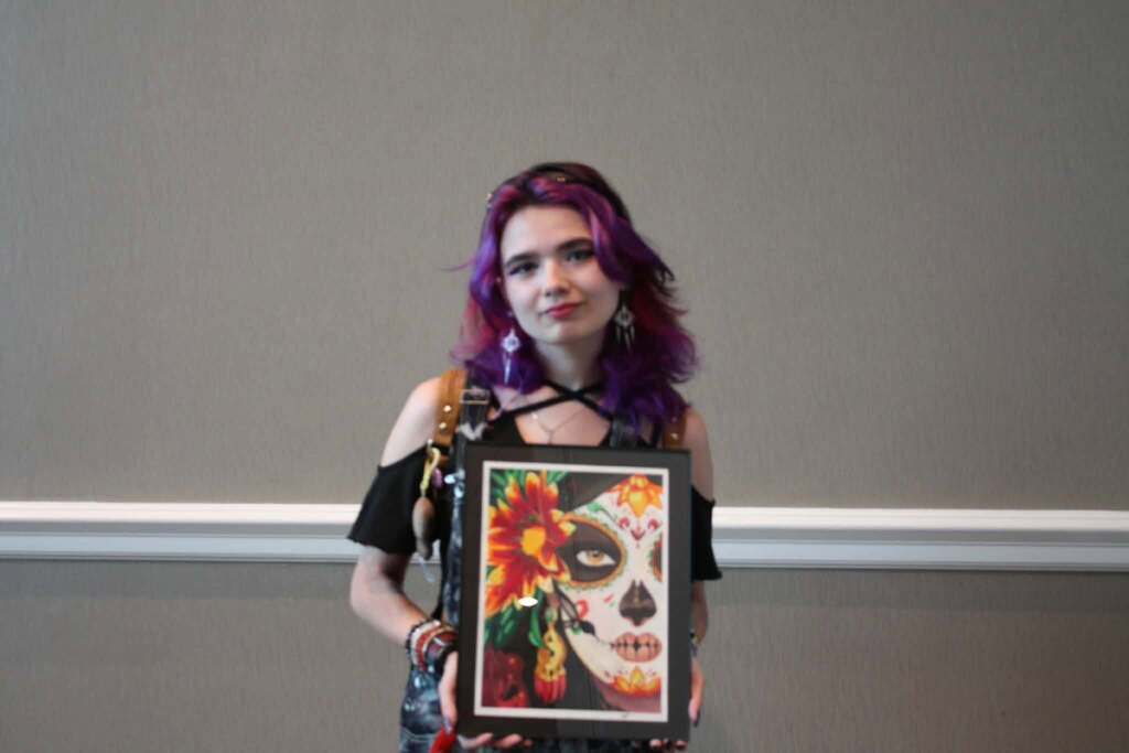 A woman with purple hair holds a framed artwork with a skull face design, standing against a gray background with a white border.