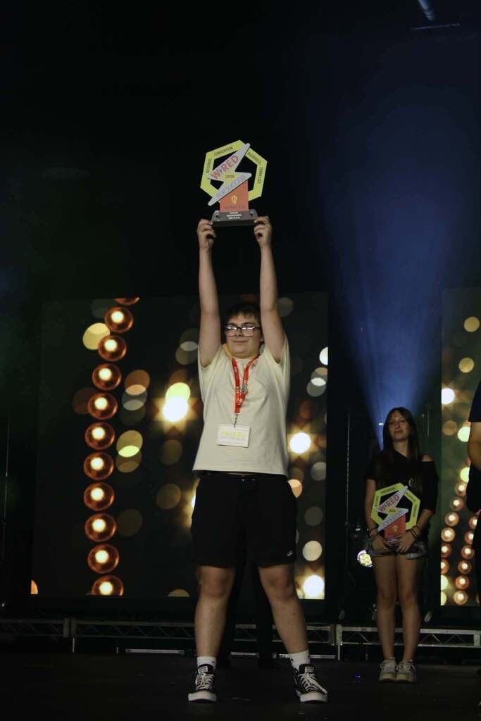 Person with a trophy raised above their head on stage, with a spotlight on them.