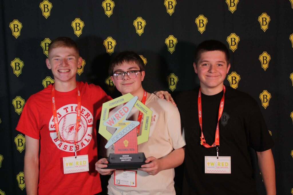 Three young men stand close, smiling, with one holding a trophy. They wear lanyards. Behind them, a black backdrop displays yellow "B" symbols.