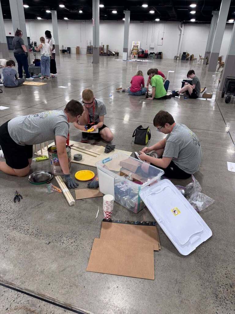 A group of individuals are gathered in a spacious room, working on a project with materials like wood and plastic on the floor.