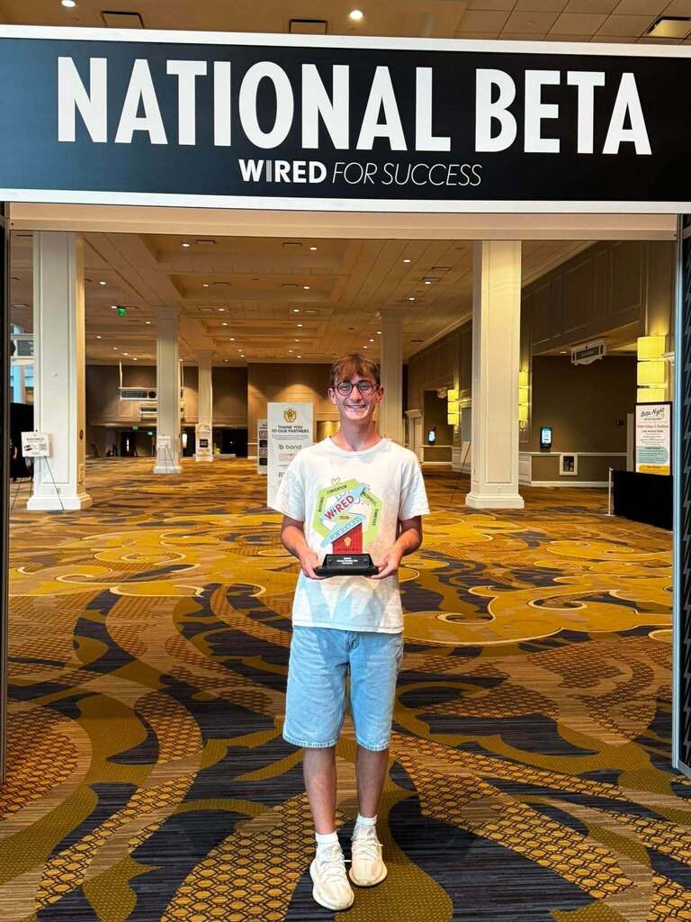 Person in white shirt and blue shorts holding a trophy stands in front of a sign that says "National Beta Wired for Success."