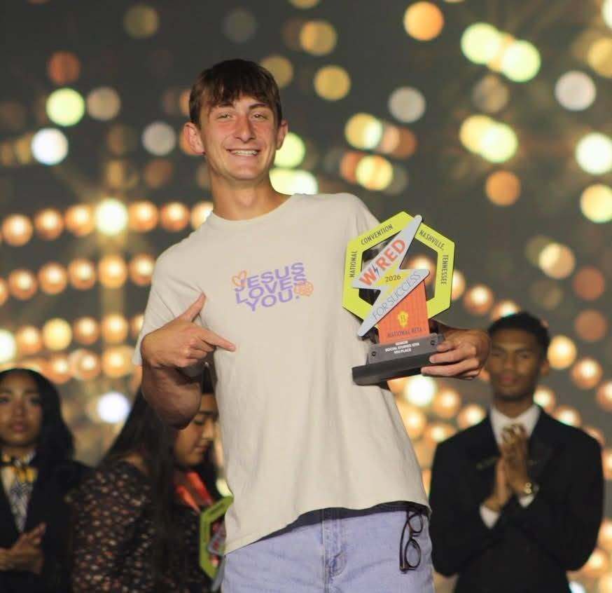 Man with trophy and thumbs up gesture standing among people at event, one clapping, others watching, with colorful lights in background.