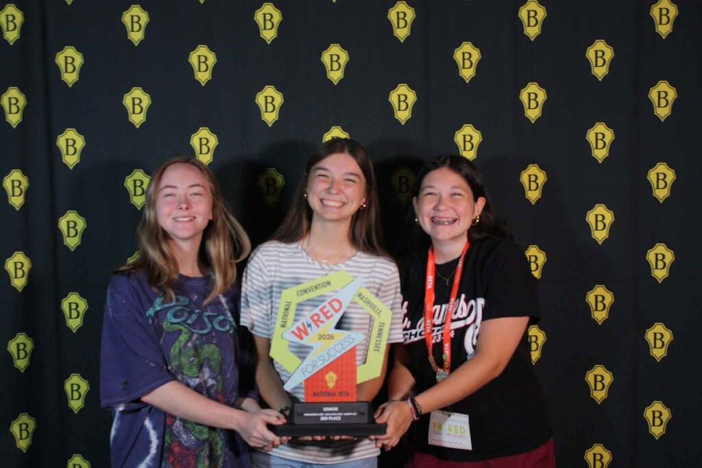 Three young people stand with a trophy. The woman in the middle holds it. They smile for the camera.