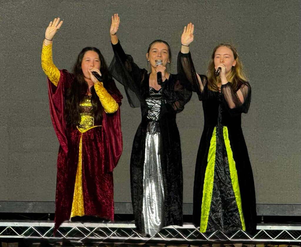 Three women on a stage, dressed in medieval-inspired costumes, holding microphones and raising their hands, possibly singing.