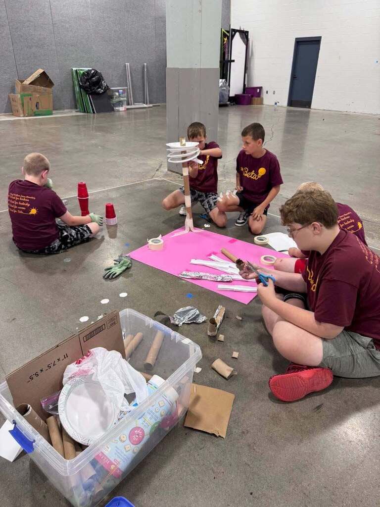 Four people are sitting on the floor in a large room, engaged in an activity. They have various materials spread out on the floor and around them.