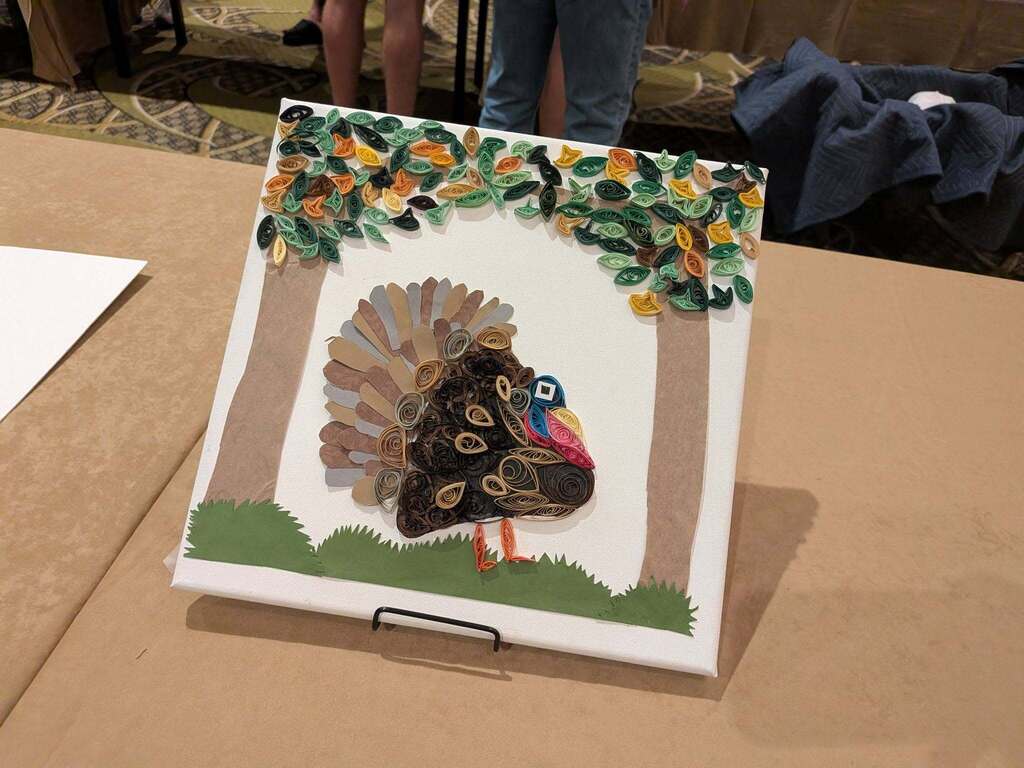 A handmade turkey art piece on a brown table. The turkey is made of colorful leaves and glass.