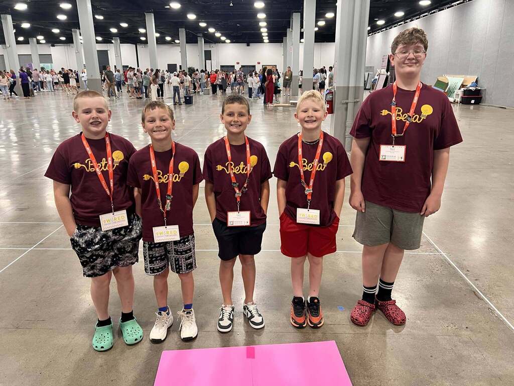 Five boys in matching shirts stand together at an indoor event. They wear ID cards and sneakers, with one wearing a watch.