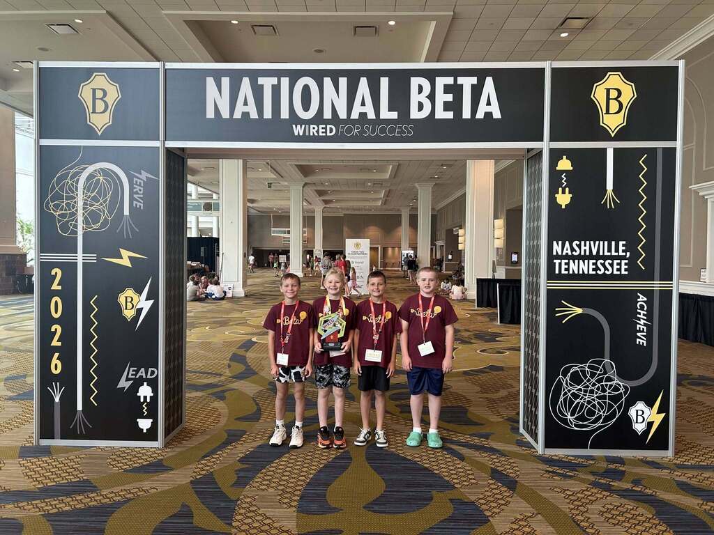 Four people in matching shirts stand under a framed banner that reads "National Beta". A geometric pattern decorates the wall behind them.