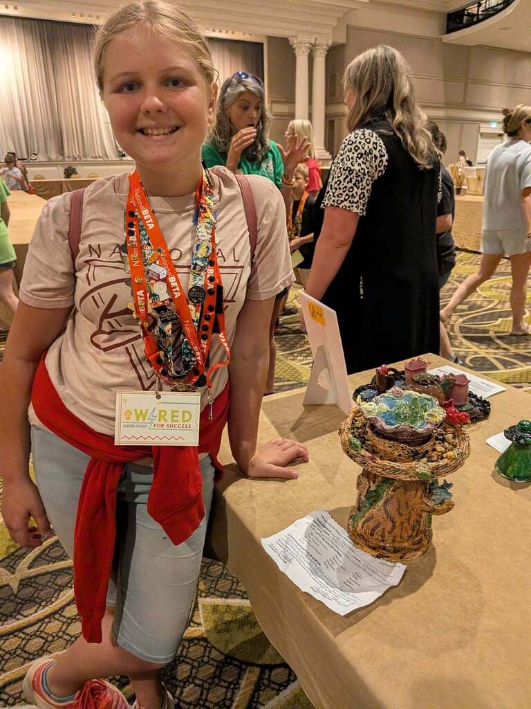 A girl with a red sash smiles at a table of art pieces in a convention center.