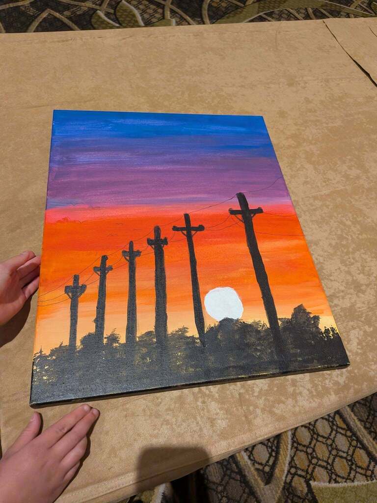 A painting of silhouettes of tall crosses on a canvas against a sunset background, placed on a table.