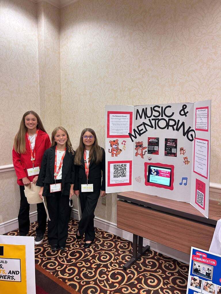 Three students stand before a music and mentoring poster with a screen and QR codes. They wear IDs and stand in a room with beige walls.